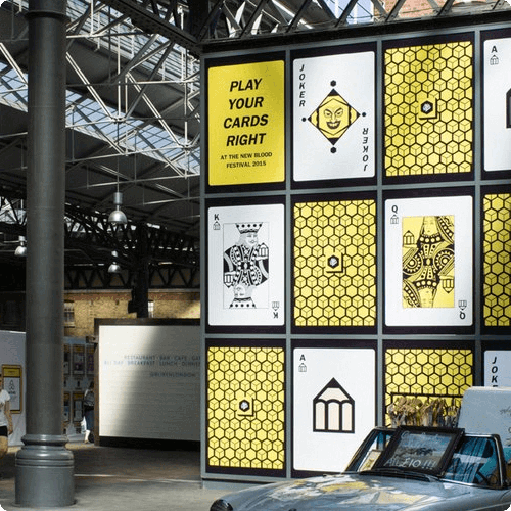 A vibrant installation featuring large, colorful playing cards and a prominent banner that reads "PLAY YOUR CARDS RIGHT" at a festival venue, surrounded by an industrial-style setting.