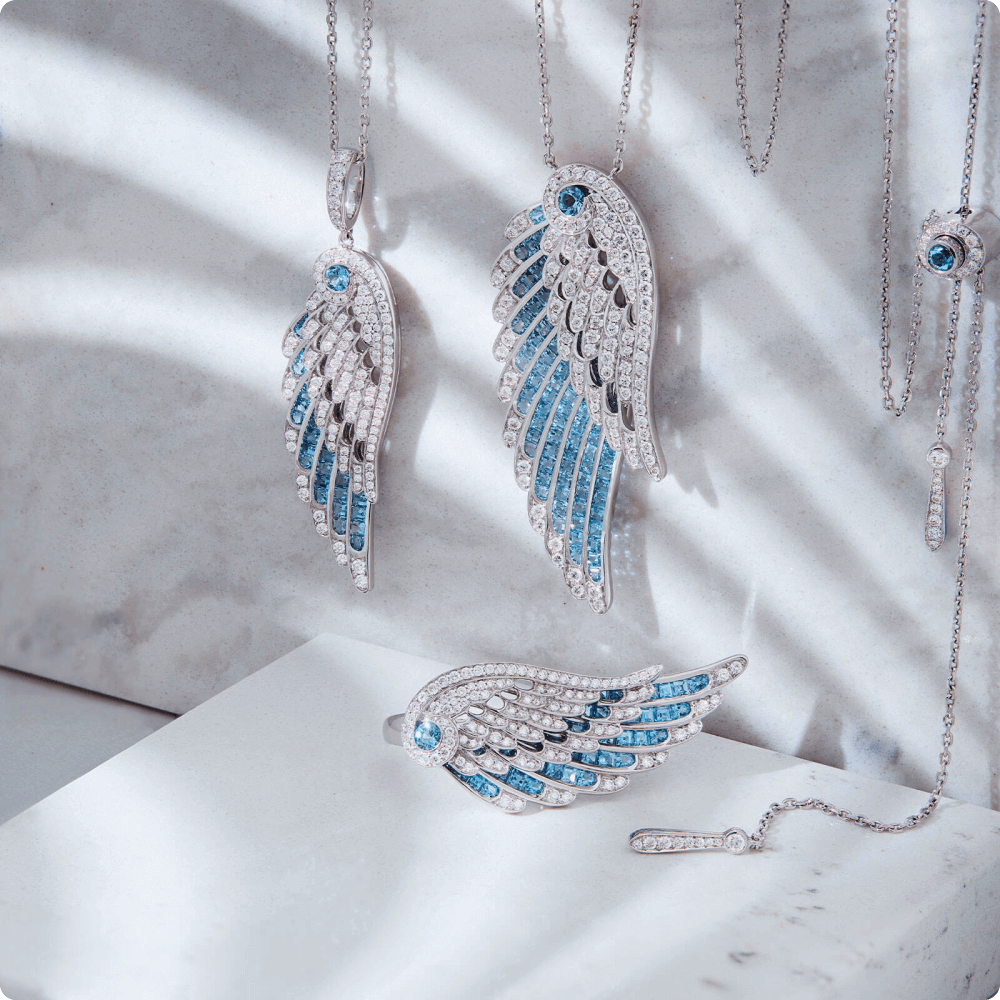 Jewelry display featuring intricate wing-shaped pendants and earrings, crafted with shimmering blue stones and silver accents. The sunlight casts delicate shadows, highlighting the elegance of each piece.