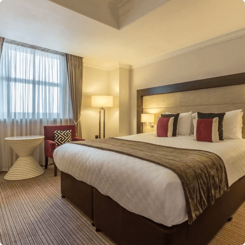 A well-appointed hotel room featuring a comfortable bed with decorative pillows, modern side chairs, a stylish lamp, and a round table, all illuminated by natural light from a nearby window.