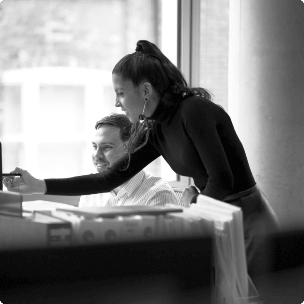 A woman stands at a desk, engaging with a man seated beside her, both focused on a digital device. The workspace features light-colored furnishings and a large window in the background, creating a collaborative atmosphere.