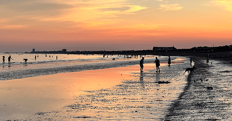 A photo of Shoreham Beach near Brighton at sunset, the sky is coloured light blue, orange and red. There are a few different silhouettes of people along the beach, throwing a ball for their dog, walking in the sea and walking along the beach.