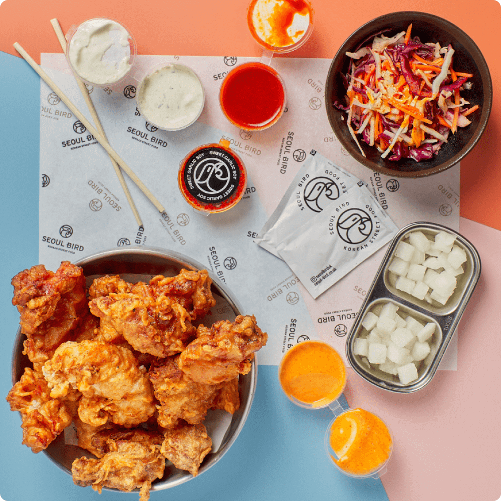 A vibrant spread featuring crispy fried chicken, colorful sauces, a fresh slaw, and sides in stylish bowls, all presented on branded paper.