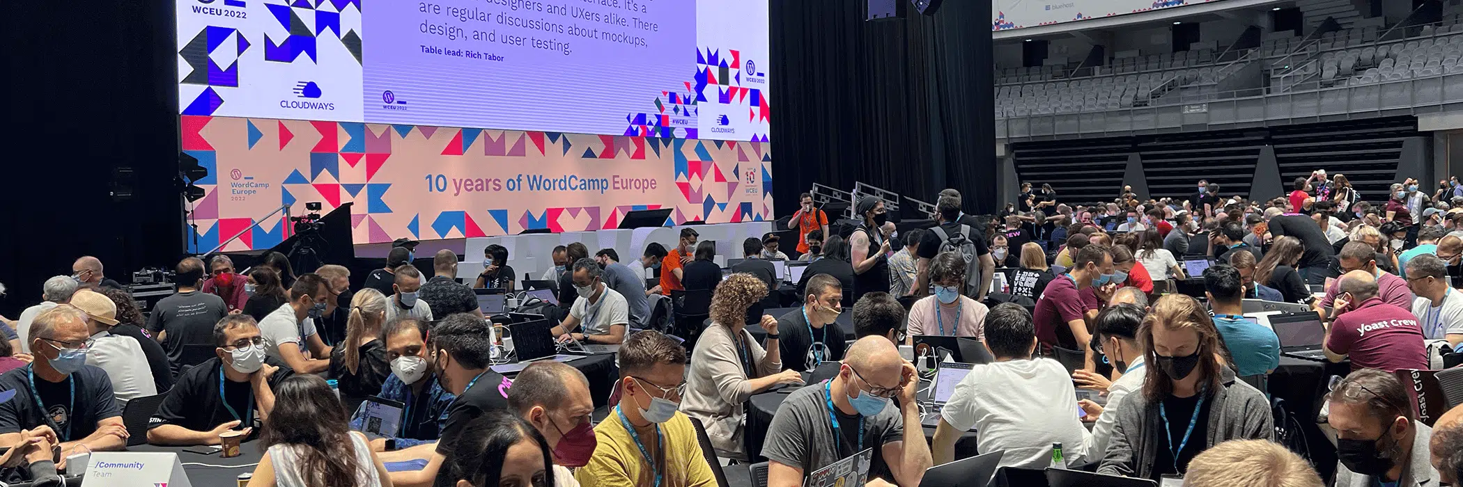 A large group of people sit at tables in a conference hall, engaged in discussions. A big screen displays text and geometric patterns in the background. Many attendees wear masks, indicating a health-conscious environment.