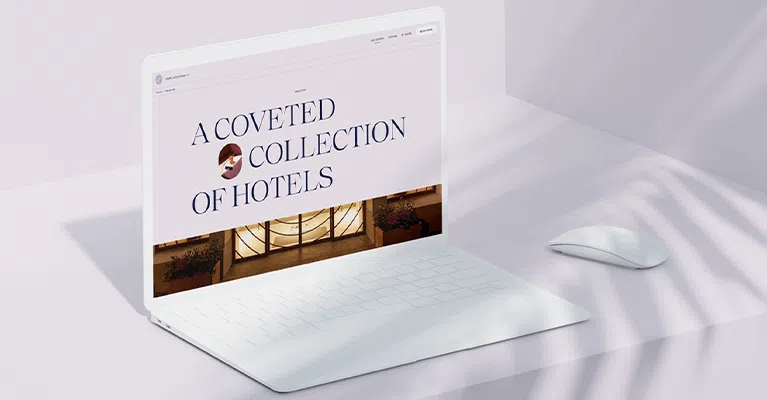 A sleek white laptop displaying a website with the text "A Coveted Collection of Hotels." There's a warm-toned image of a building interior below the text. A white wireless mouse is placed beside the laptop on a light surface with soft shadows.