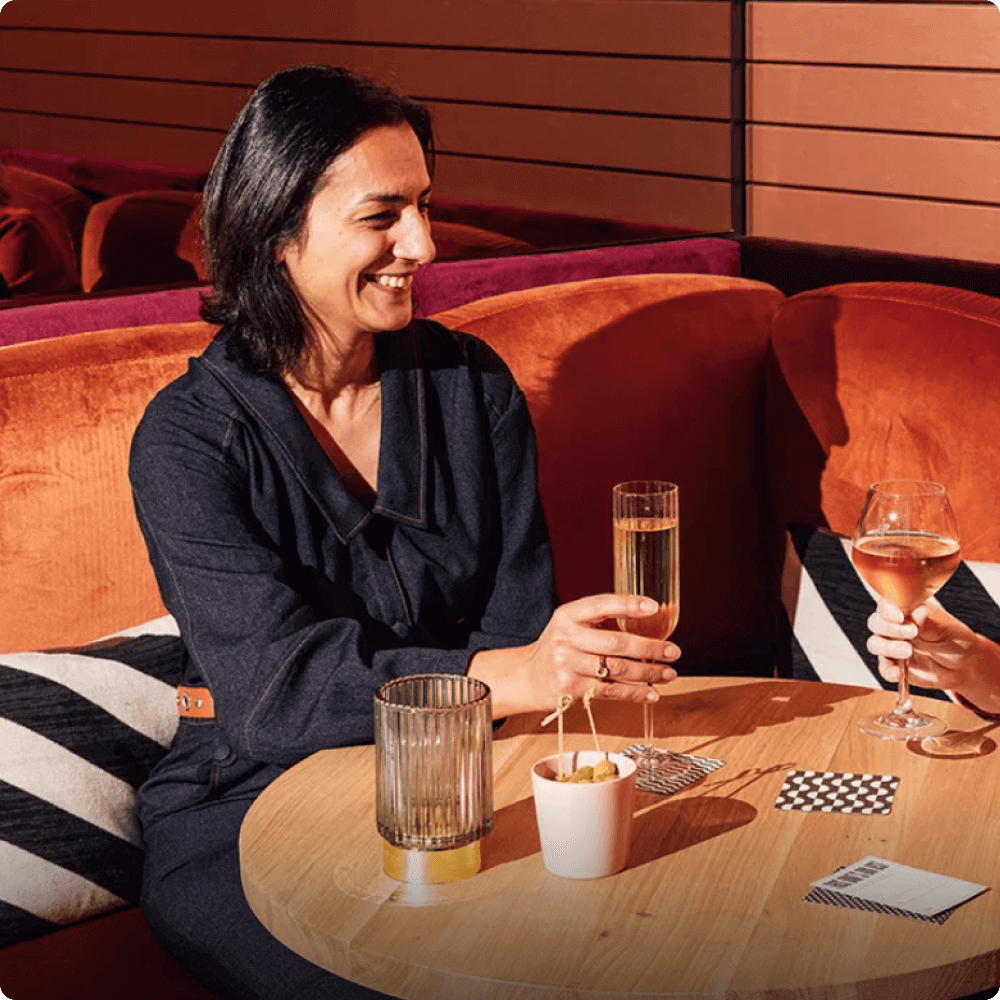 A woman smiles while holding a glass of champagne, seated at a table with another person. The setting features warm colors and stylish furnishings, creating a relaxed atmosphere for conversation and enjoyment.