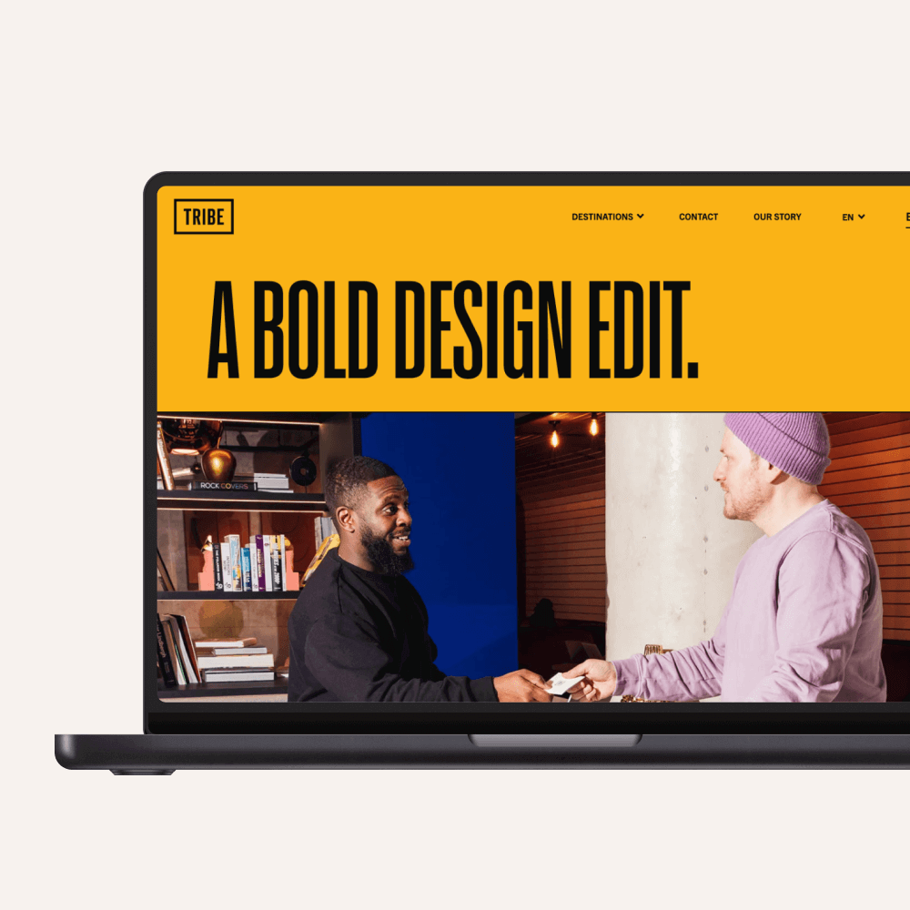 A laptop displaying a vibrant website featuring two men engaging in conversation. The bold yellow header reads "A BOLD DESIGN EDIT." The background includes shelves with books, suggesting a creative environment.