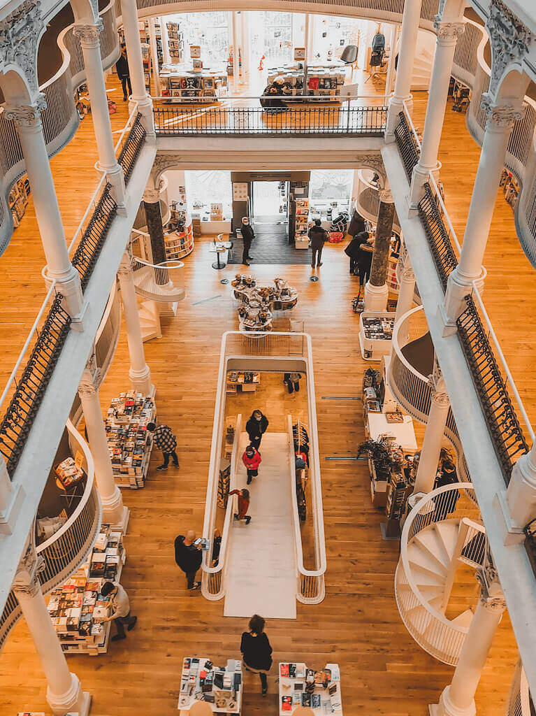Aerial view of a multi-level bookstore with wooden floors and spiral staircases. People browse shelves filled with books, and natural light streams in through large windows, creating an inviting atmosphere.