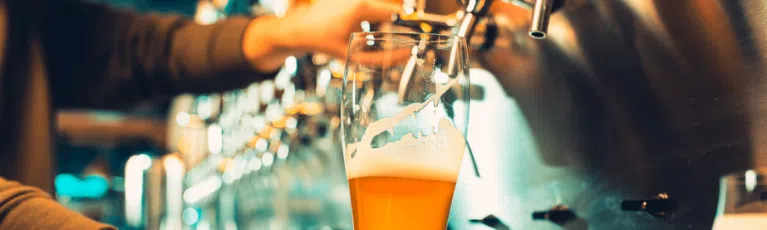 A bartender pours a glass of amber beer from a row of shiny metal taps, capturing the frothy head. The image highlights the action of serving beer in a bar setting.