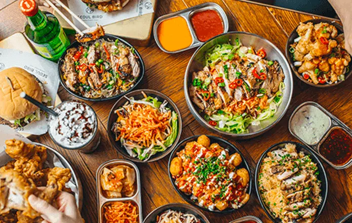 An overhead view of a table filled with various dishes including salads, fried chicken, a burger, sauces, and drinks. The vibrant array of foods showcases colorful vegetables, grilled meats, and flavorful garnishes, suggesting a feast or meal gathering.