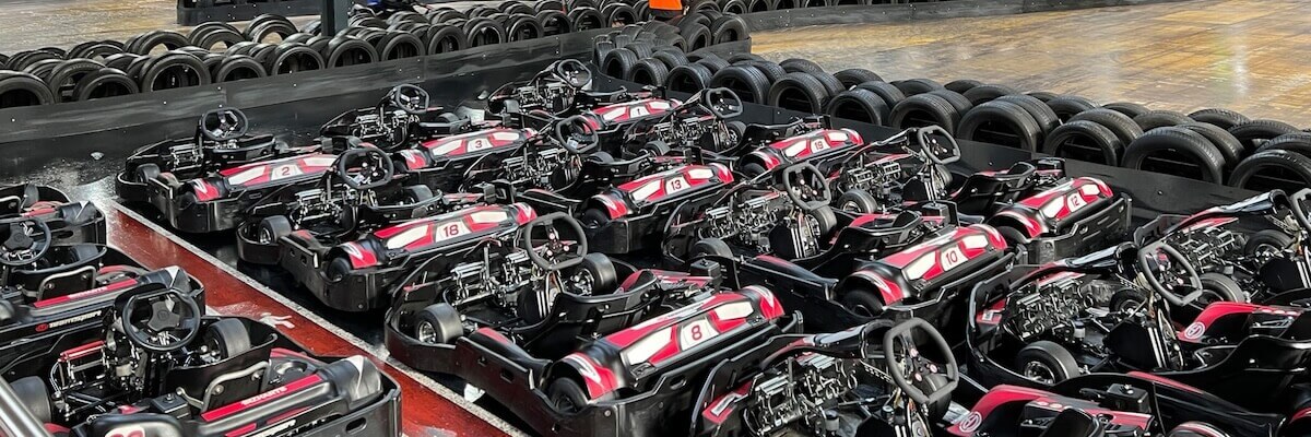 Rows of parked go-karts in an indoor racing track, surrounded by black protective barriers. Each kart is black with red and white accents, neatly aligned, ready for racing.