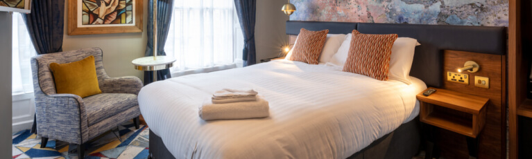 A cozy hotel room featuring a neatly made double bed with white linens, two orange pillows, and folded towels. An armchair with a yellow cushion sits by a window, alongside a small round table. The room is decorated with a large framed artwork and dark curtains.