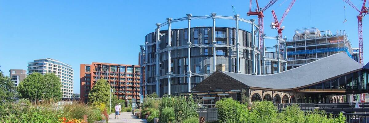 Urban scene with modern buildings and construction cranes under a clear blue sky. The foreground has lush greenery and a walking path, while a cylindrical building and other structures rise in the background.