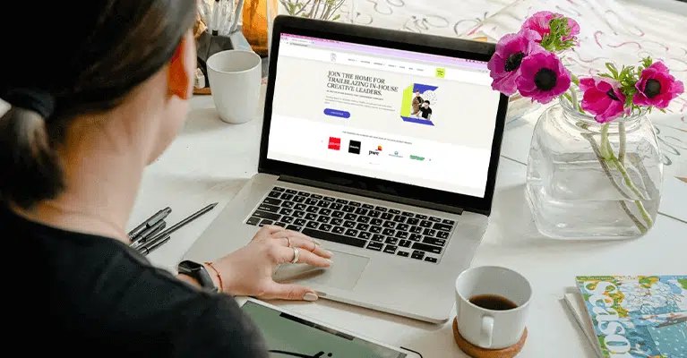 A person sits at a desk, browsing a website on a laptop. The desk holds a cup of coffee, a magazine, and a vase with pink flowers. The scene suggests a casual, work-from-home setting.