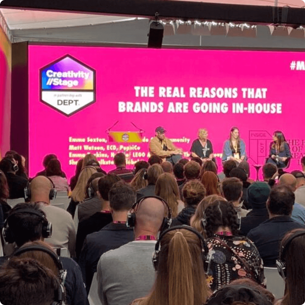 A panel discussion featuring industry experts on the reasons brands are transitioning to in-house operations. The backdrop displays a bright pink screen with the title of the talk, while the audience listens attentively.