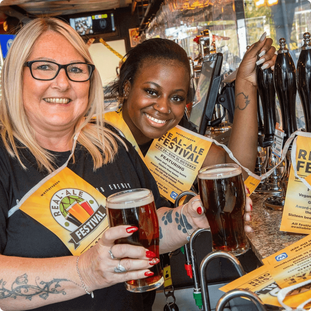 Two smiling women hold pints of beer at a bar, both wearing festival lanyards. Behind them, beer taps and festival promotional materials are visible, creating a lively atmosphere celebrating a beer festival.