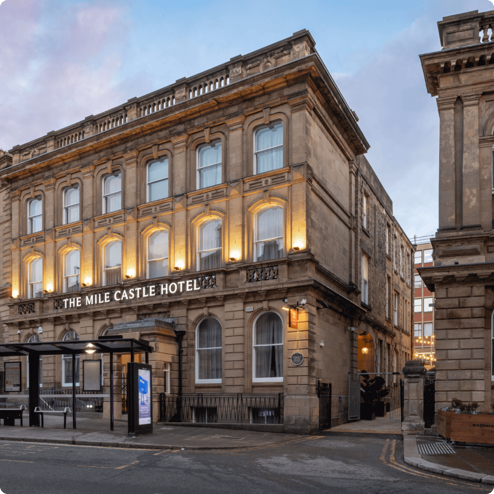 The image shows the exterior of The Mile Castle Hotel, featuring classic architecture with illuminated windows. The hotel is situated on a street with a bus stop nearby, emphasizing its convenient location.