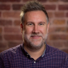 A person with short gray hair and a beard is smiling, standing in front of a brick wall. They are wearing a checkered shirt.
