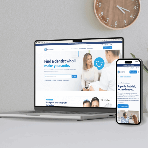 A laptop and smartphone displaying a dental appointment website with "Find a dentist who'll make you smile" text. Both devices sit on a white surface with a wooden wall clock visible in the background.