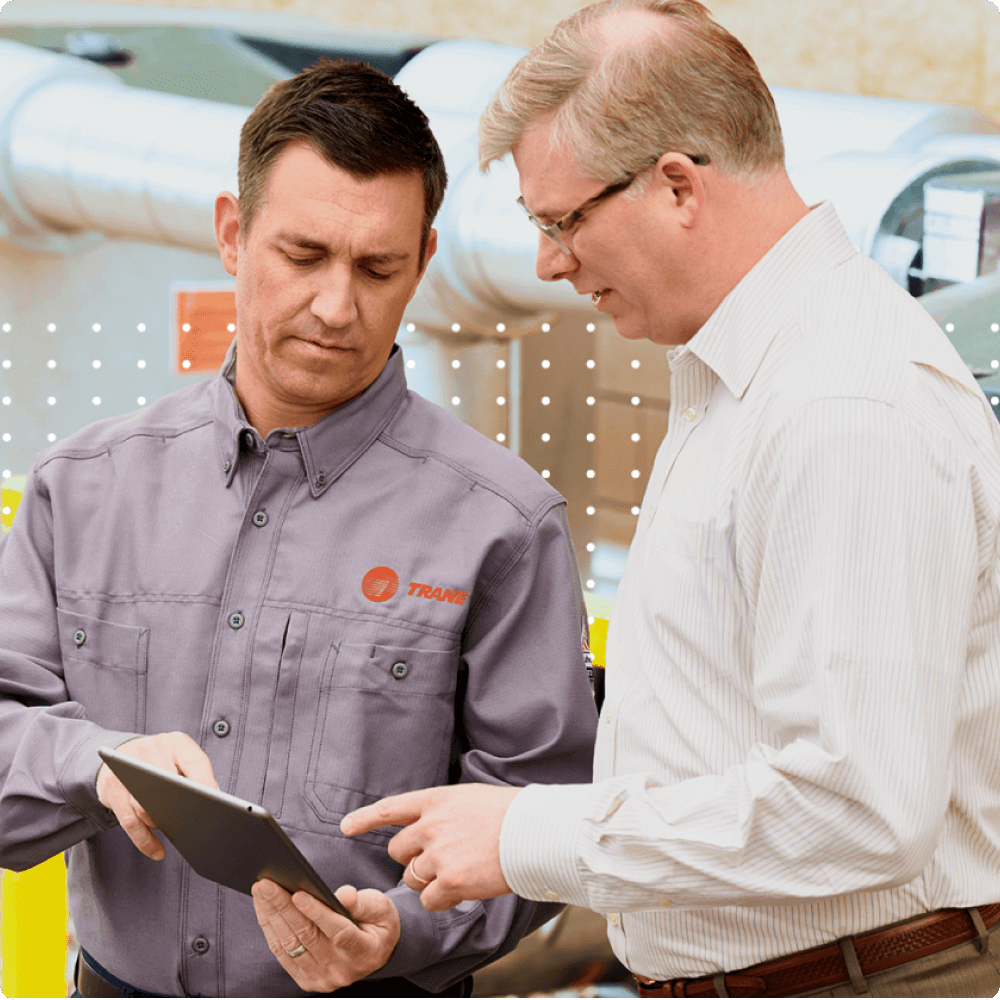 Two professionals engage in a discussion, reviewing information on a tablet. One is in a gray shirt, while the other wears a white dress shirt. They appear focused and collaborative in a workplace setting.