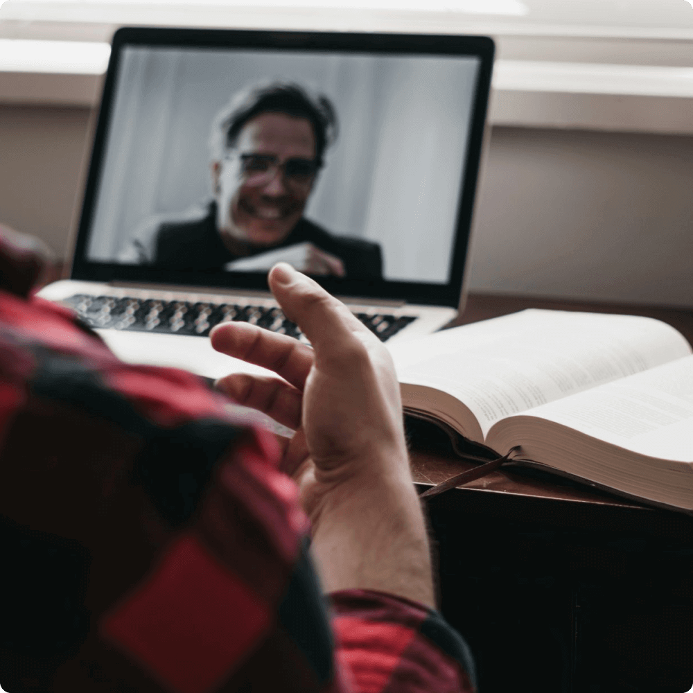 A person sits at a desk, engaged in a video call on a laptop. The screen displays a smiling individual, while a large, open book rests nearby. The setting suggests a collaborative learning or discussion environment.
