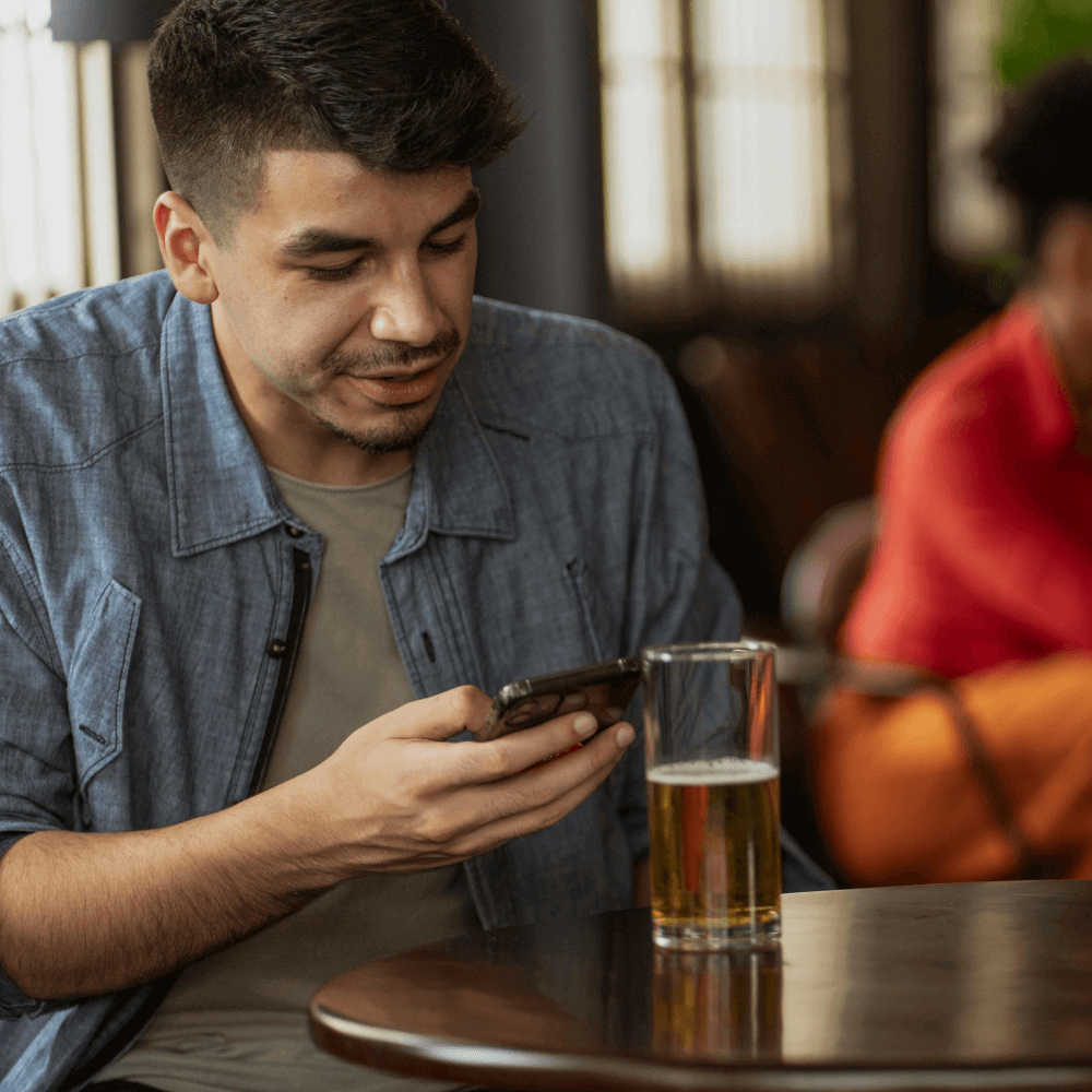 A man sits at a table in a casual setting, looking at his smartphone. A glass of beer rests beside him. Another person can be seen in the background.
