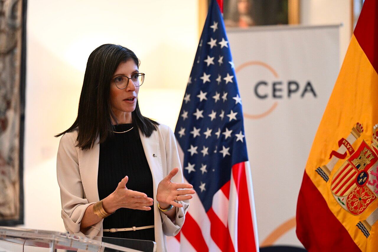 A woman speaks at a podium with U.S. and Spanish flags in the background, discussing topics related to international collaboration. The setting suggests a formal event, possibly focused on diplomacy or trade.