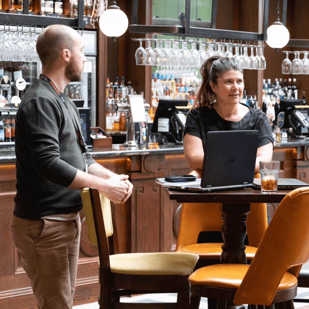 A man and a woman engage in conversation at a bar. The woman sits at a table with a laptop, while the man stands nearby. A variety of drinks and a well-stocked bar are visible in the background.