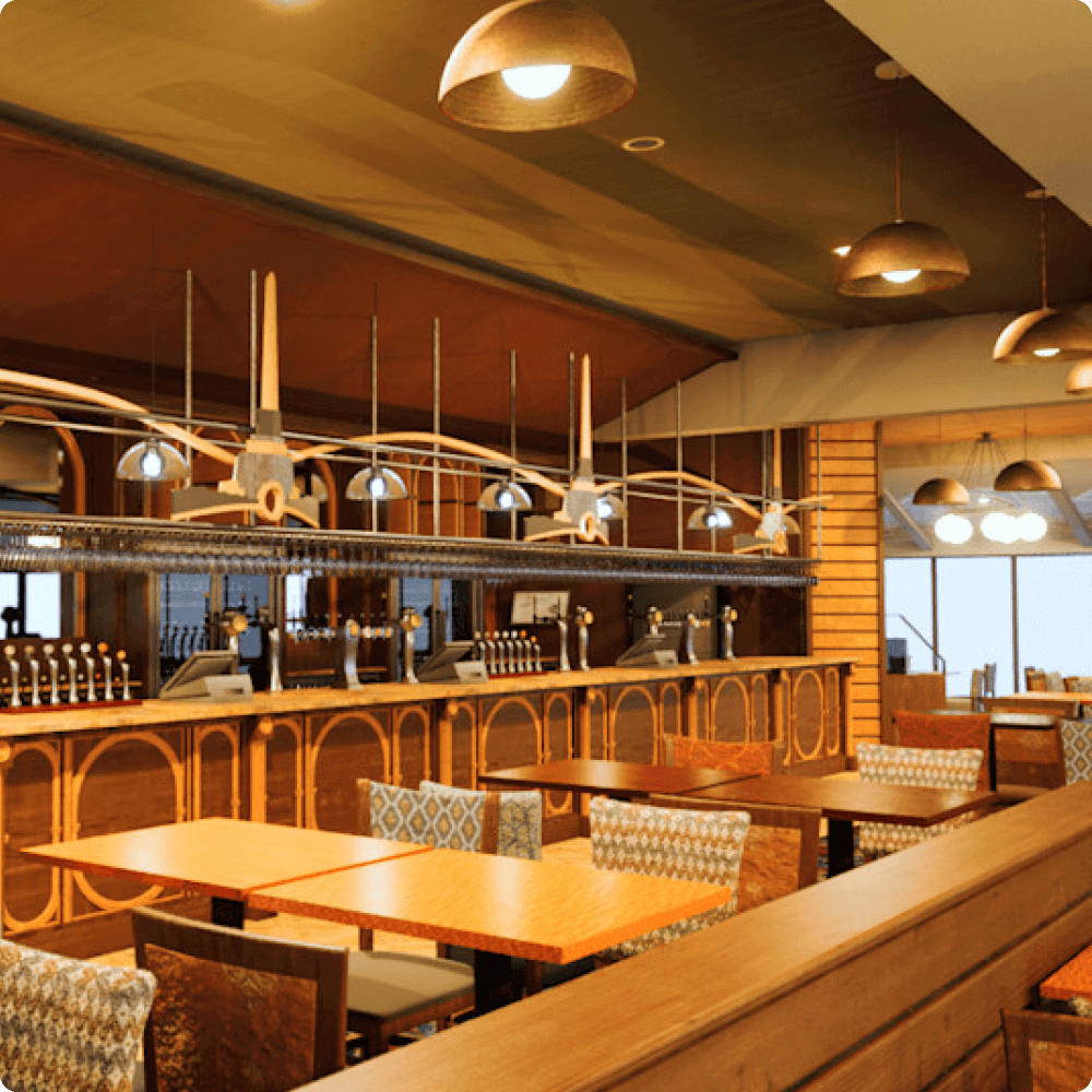 A modern restaurant interior featuring wooden tables and chairs, pendant lighting, and a bar area. The design creates a warm, inviting atmosphere for diners.