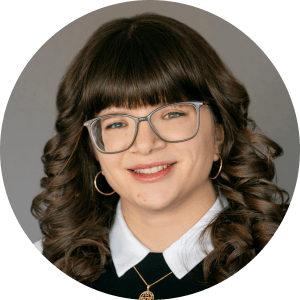 A person with long, curly brown hair, wearing glasses and a black top with a white collar. They are smiling against a plain background.