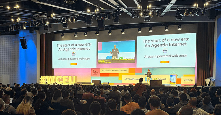 A speaker presents at a conference in a large auditorium with a slide titled "The start of a new era: An Agentic Internet." The audience is seated, and stage lights illuminate the scene.