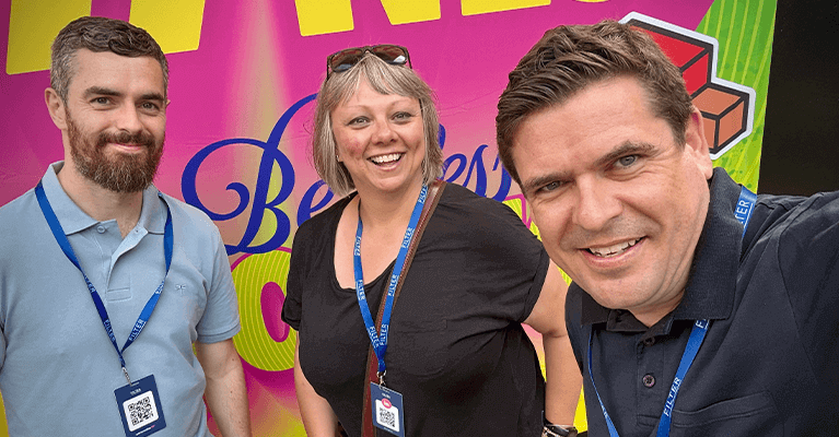 Three people smiling and wearing lanyards stand in front of a colorful background with text and geometric shapes.