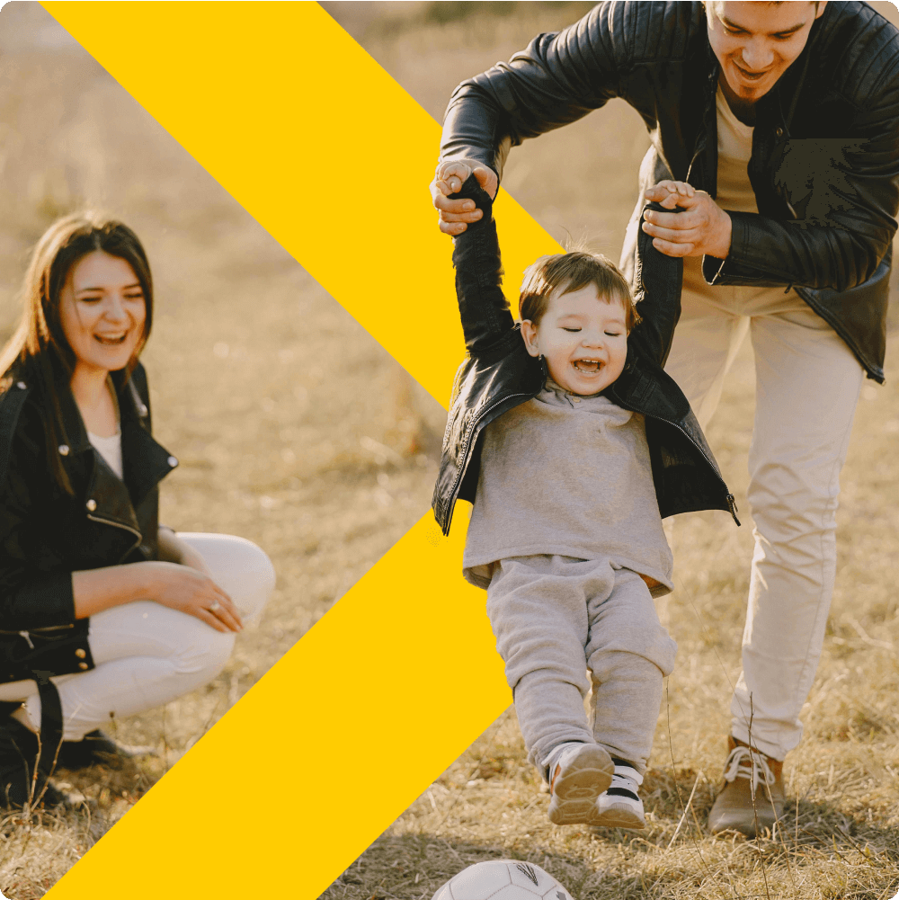 A smiling child joyfully hangs from their parent's hands in an outdoor setting, while another adult, seated nearby, watches with a smile. The background features a yellow diagonal design, adding a vibrant touch to the scene.