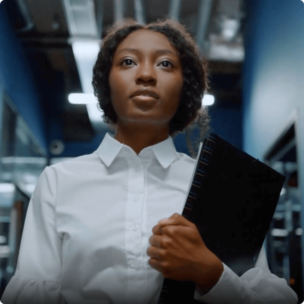 A confident woman in a white shirt stands in a modern workspace, holding a notebook. The background features shelves and a well-lit corridor, suggesting a professional environment focused on collaboration and productivity.