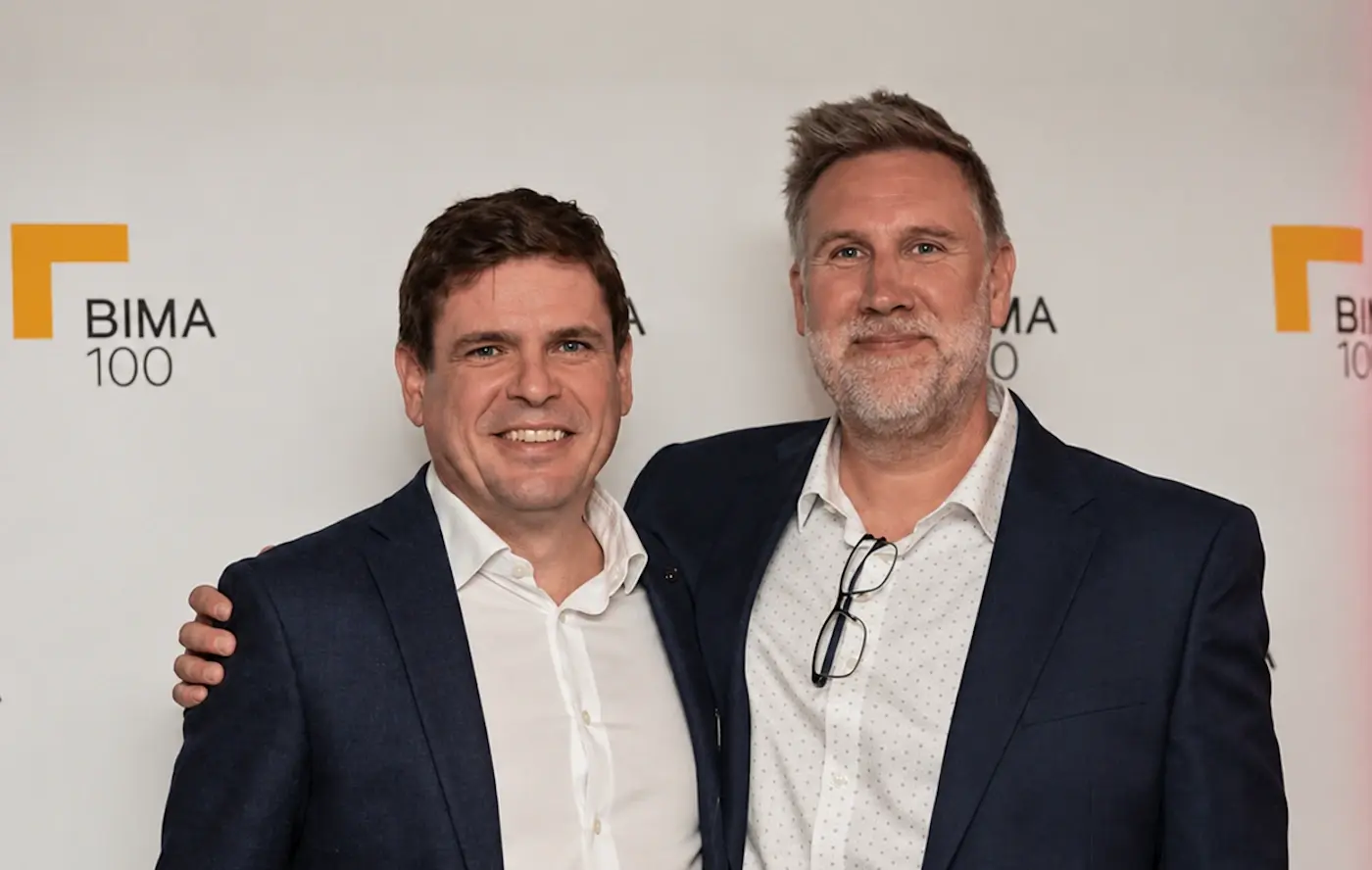 Two men in navy blazers and white open-collared shirts stand side by side in front of a BIMA 100 backdrop, smiling at the camera. One has short dark hair. The other has a grey beard, glasses hooked on his shirt, and an arm around his companion's shoulder.