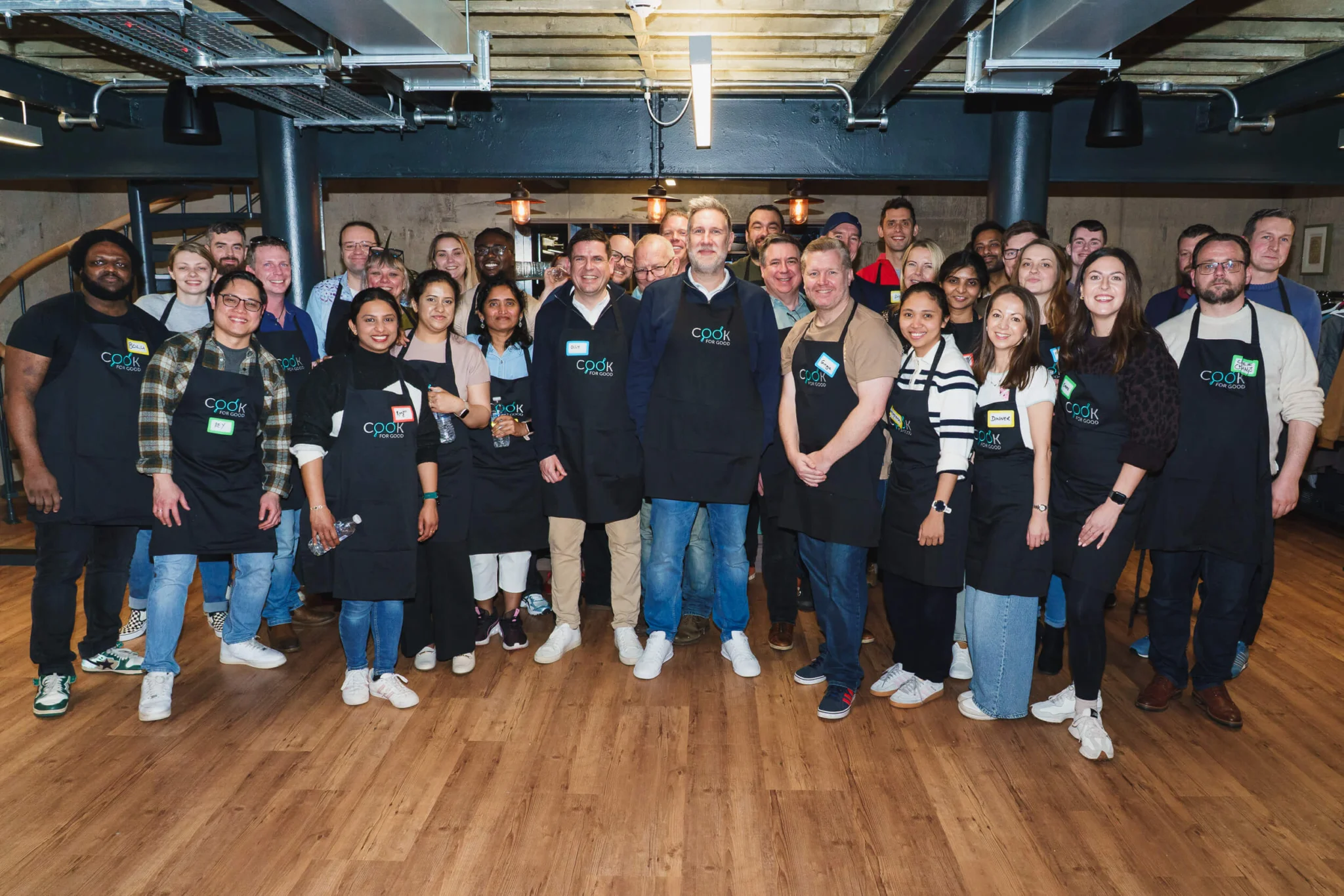 A large team of around thirty colleagues stands together in an industrial-style kitchen space with exposed pipes and wooden flooring. Most wear black "Cook for Good" aprons with handwritten name badges, smiling at the camera during a group volunteering event.