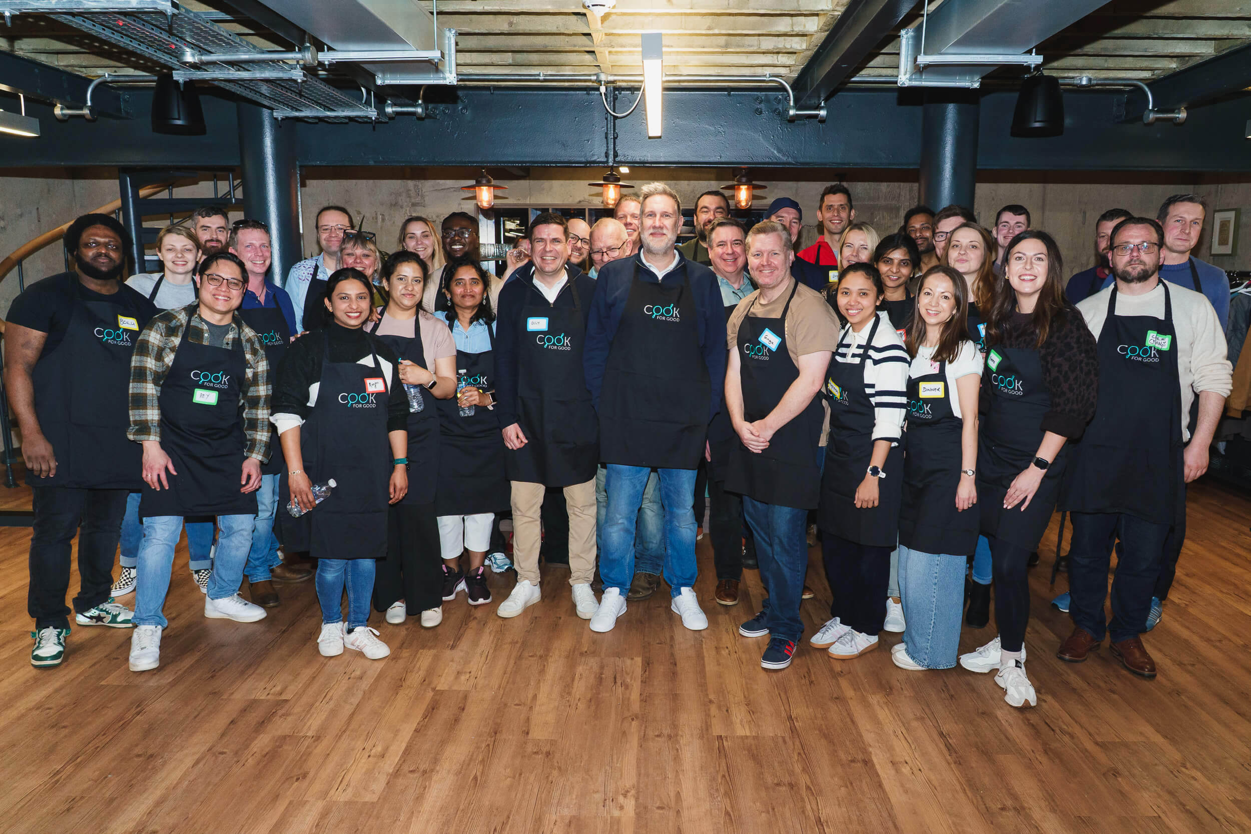 A large team of around thirty colleagues stands together in an industrial-style kitchen space with exposed pipes and wooden flooring. Most wear black "Cook for Good" aprons with handwritten name badges, smiling at the camera during a group volunteering event.