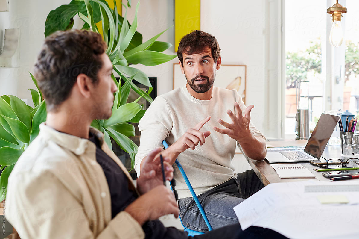 Two men talk at a desk in a bright, plant-filled office. One gestures with both hands while explaining a point — the other listens, pencil in hand. A laptop, notebook, glasses and papers sit between them. Working session, not a presentation.