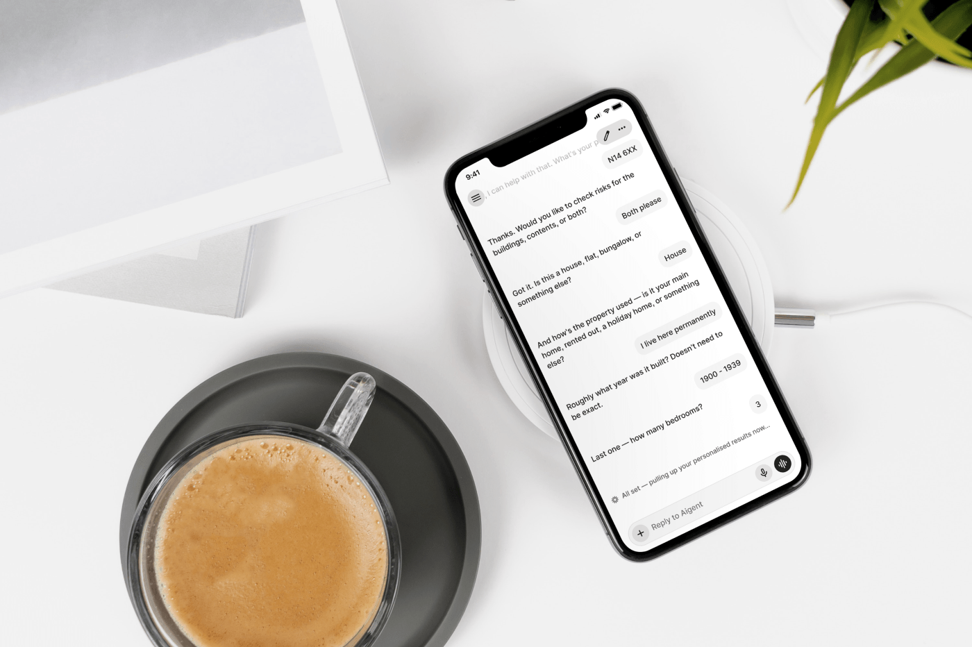 Smartphone on a wireless charger showing a chat with an AI insurance assistant. It asks about buildings and contents cover, property type, use, build year and bedrooms — the user answers in short replies. Beside it, a glass of coffee on a grey saucer, a plant and a folder on a white desk.