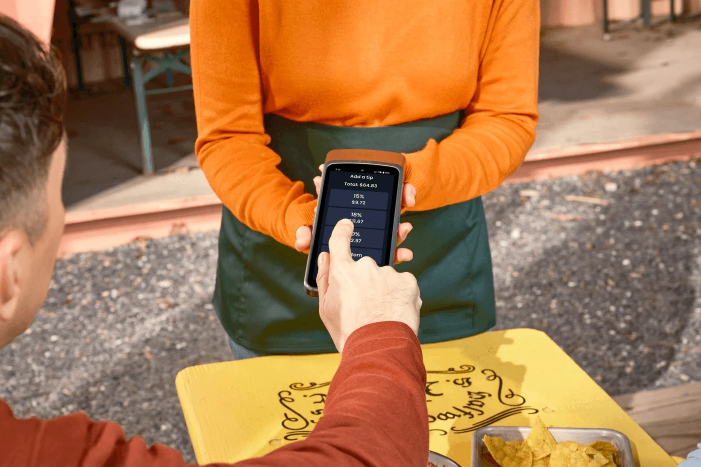 A server in an orange sweater and green apron holds a handheld payment terminal at an outdoor table. The screen shows a $64.83 total with tip options — 15%, 18%, 20% and custom. A seated customer taps 18%. A tray of tortilla chips sits nearby.
