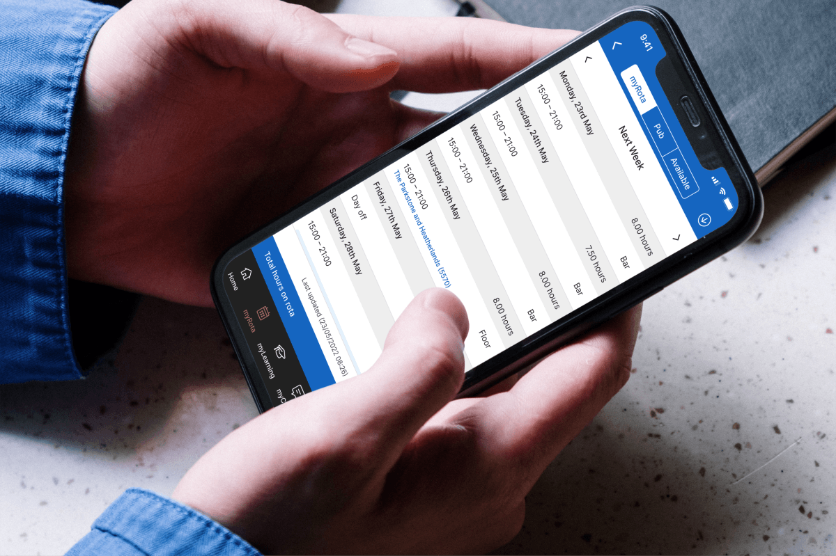 Hands in a blue denim shirt hold a smartphone showing a staff rota app. The screen lists next week's shifts by day, with times, hours and roles such as Bar and Floor. Tabs read myRota, Pub and Available. Navigation icons sit along the bottom.