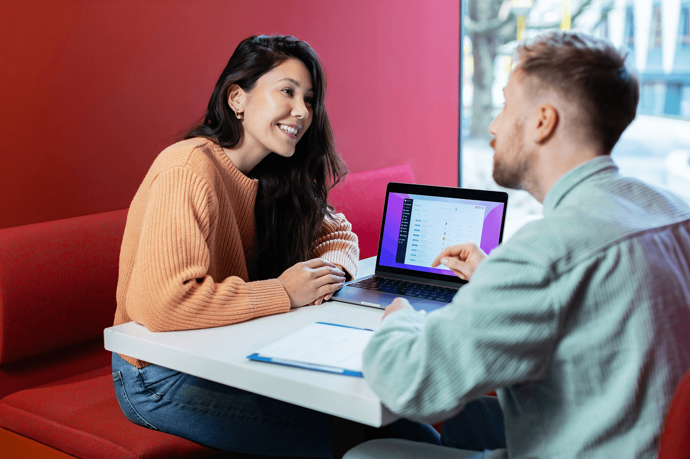 Two colleagues sit at a white table in a red office booth. A man points at a laptop screen showing a project dashboard while a woman in an orange sweater listens and smiles. A clipboard with notes lies between them. Large windows behind show trees outside.