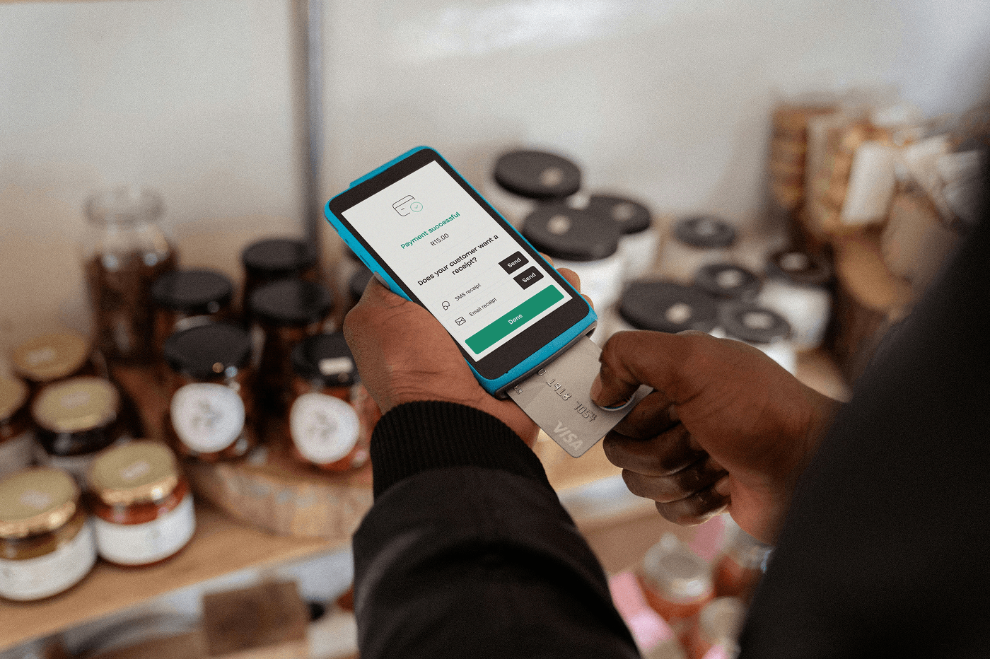 Hands hold a blue card terminal with a Visa card inserted. The screen reads "Payment successful, R15.00" and offers SMS or email receipt options. Shelves of jarred preserves and spices sit in the blurred background of a small shop.