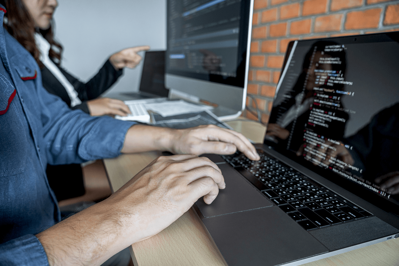 A developer in a denim shirt types on a laptop showing lines of code. Behind them, a colleague points at a monitor displaying more code. The shared desk sits against an exposed brick wall. Two engineers, focused work, not a staged pitch.
