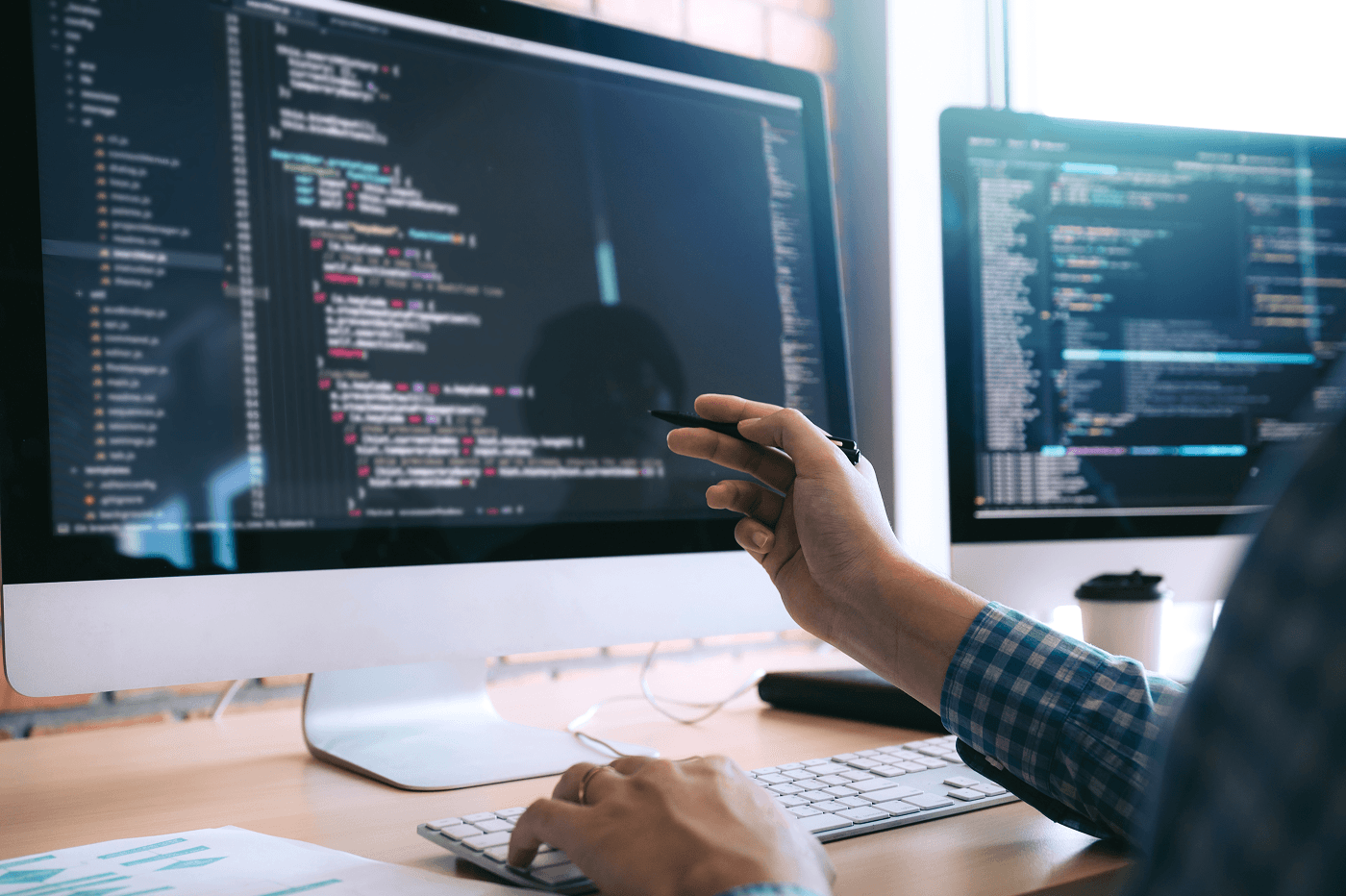 A developer in a checked shirt points a pen at lines of code on a desktop monitor, one hand on the keyboard. A second screen beside it shows more code. The desk sits near a bright window. The work is focused and hands-on, not staged.