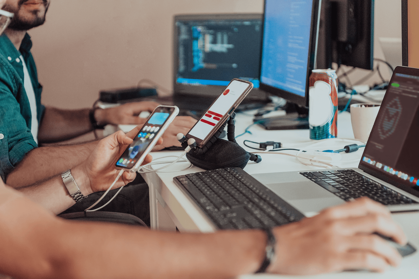 Two developers test a mobile app at a shared desk. One holds a smartphone showing the app, while another phone sits on a stand beside a laptop. Monitors displaying code fill the background, with cables and a drink can on the desk.