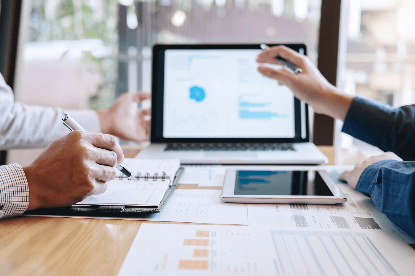 Two colleagues review business data at a wooden desk. One points a pen at a laptop displaying blue charts, the other takes notes in a spiral notebook. A tablet and printed spreadsheets with bar graphs sit between them — a working session, not a presentation.